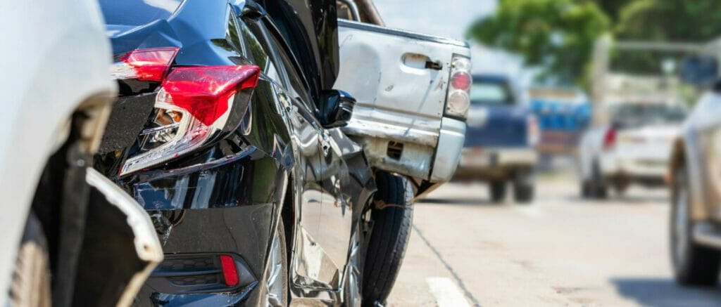 A close-up, ground-level shot of a multi-car pileup on a highway, showing heavy damage to the rear of a black sedan and a silver pickup truck.