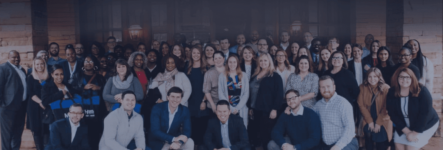 A wide, panoramic group photo of a large, diverse corporate team standing together in front of a stone building.