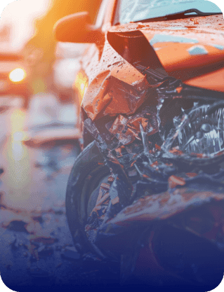 A close-up of a badly damaged orange car after a collision, with crushed metal and a shattered headlight visible against a sunset background.