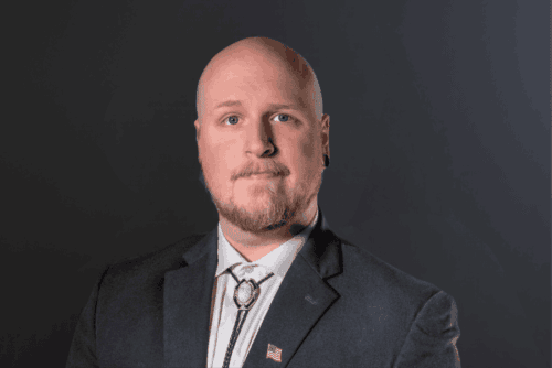 Portrait of a bald man with a light beard in a dark suit and white shirt, wearing a bolo tie, against a dark gradient background.